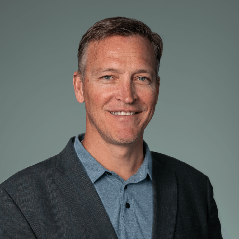 Professional headshot of Kevin Crawley, smiling and wearing a dark blazer over a blue shirt, posed against a light gray background.