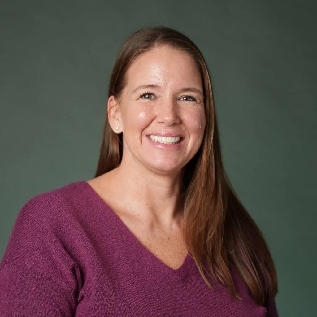 Professional headshot of Heather Beckner, smiling and wearing a purple top, posed against a neutral gray background.